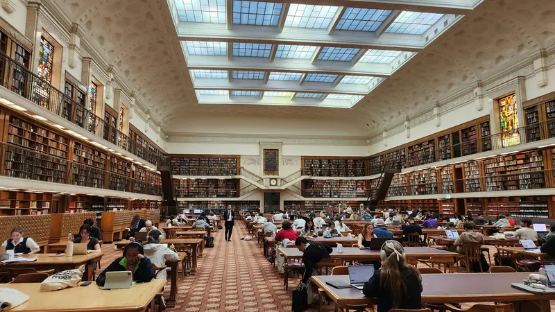 Library reading tables surrounded by bookshelves beneath broad ceiling windows